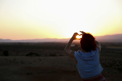 Rear view of woman gesturing heart shape against sky during sunset