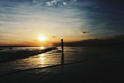 Silhouette boy standing at sea shore against sky during sunset