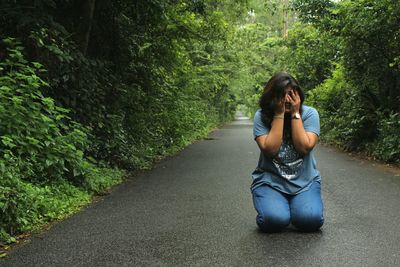 Young woman hiding face while kneeling on road amidst trees