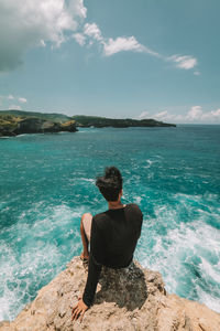 Rear view of man sitting on rock by sea against sky