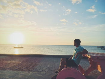 Man sitting at beach against sky during sunset