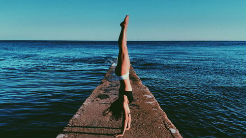 Side view of young woman practicing handstand on pier over sea against sky