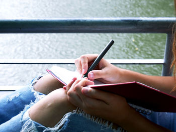 Midsection of woman writing in book while sitting against lake