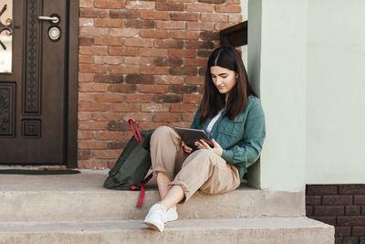 Young female student sitting on steps and studying with tablet in college or private school