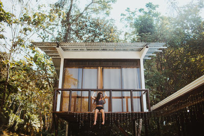 From below full body of young long haired female traveler in stylish swimwear sitting on balcony of beach house located near green trees in summer day in uvita town in costa rica