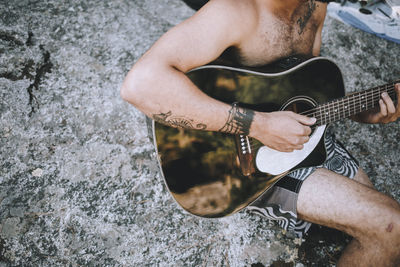 Midsection of shirtless man playing guitar on rock