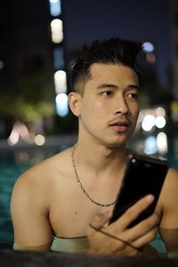 Thoughtful shirtless young man holding smart phone while looking away in swimming pool at night