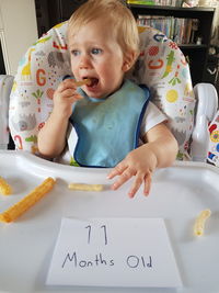 Cute baby boy eating food while sitting on high chair with message at home