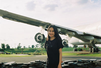 Portrait of smiling young woman standing by airplane against sky