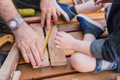 Crop unrecognizable dad measuring wooden piece with tape against child with pencil sitting on boardwalk