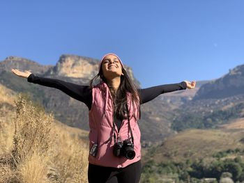 Portrait of young woman standing against sky in mountains, open arms and free