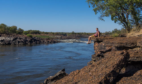 Man sitting on rock over river against clear sky