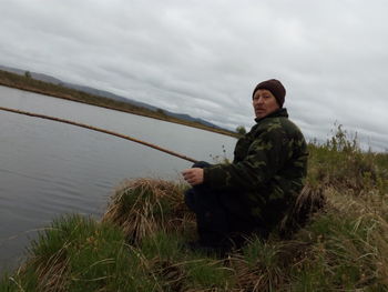 Portrait of mature man fishing in lake while sitting on grassy field against cloudy sky