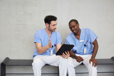 Male nurses discussing over digital tablet while sitting against gray wall in corridor at hospital