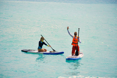 Young man dressed in swimsuit and lifevest waving and young woman paddling on stand up paddle board