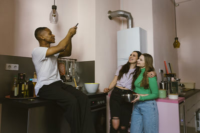 Smiling man sitting on counter photographing young friends in kitchen at home