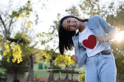Smiling young woman standing by heart shape against trees