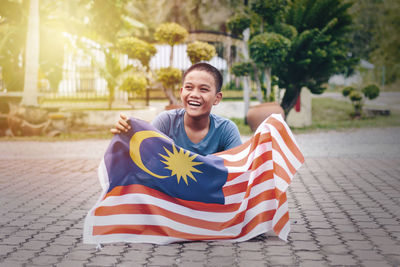 Smiling boy holding malaysian flag while sitting on footpath
