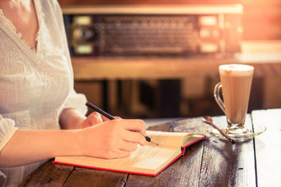 Midsection of young woman writing on book at wooden table