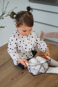 A little girl sits on a wooden table next to a metal basket with easter eggs. 