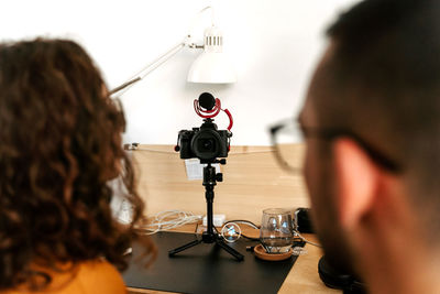 Back view of focused young couple blogger with curly hair in casual outfit and eyeglasses and professional photo camera placed on tripod while shooting video at home