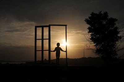 Silhouette boy walking at gate on field against sky during sunset