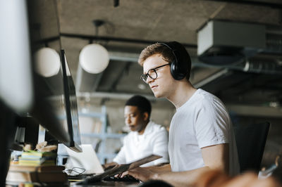Young male programmer doing coding on computer with wireless headphones in office