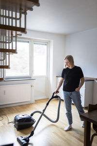 Full length of young woman using vacuum cleaner on hardwood floor at home