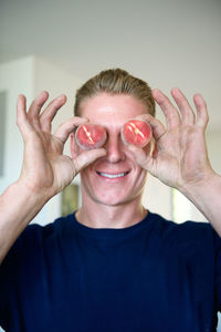 Smiling young man holding peaches in front of eyes at home