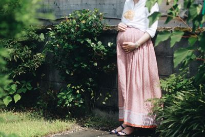 Low section of pregnant woman standing by surrounding wall in yard