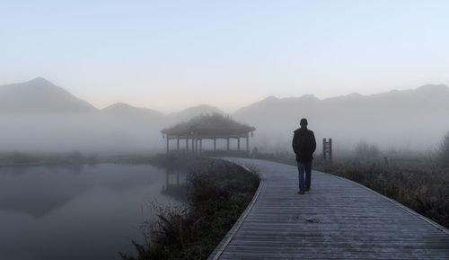 Rear view of man walking on pier over lake against clear sky during foggy weather