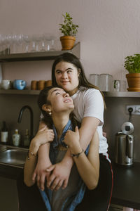 Young woman with down syndrome hugging sister from behind in kitchen at home