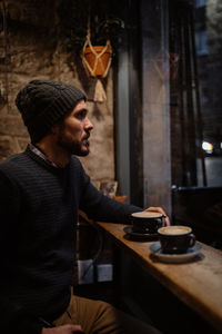 Side view of tranquil male in warm sweater and hat sitting at counter in cozy wooden cafe and enjoying hot delicious coffee while looking away