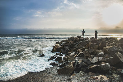 People standing on rocks at beach against sky during sunset