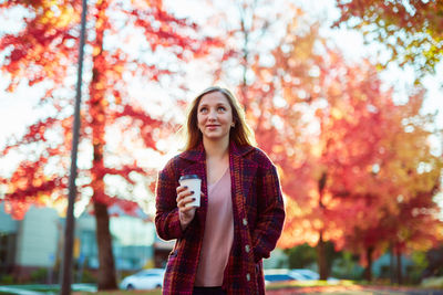 Young woman holding coffee while standing against autumn trees