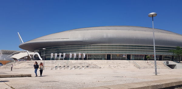 People walking by modern building against clear blue sky