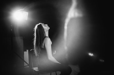 Young female model looking up while sitting on chair by illuminated light in studio