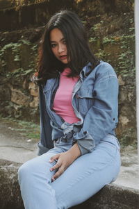 Portrait of young woman wearing denim jacket while sitting on retaining wall