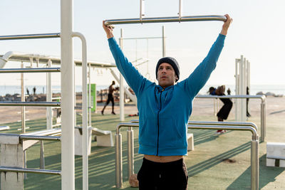 Active ethnic male in warm sportswear hanging on pull ups bar while training on sports ground with fitness equipment