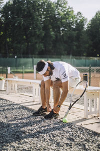 Man tying shoe lace while sitting on bench at tennis court