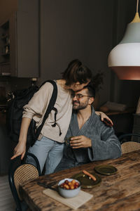 Non-binary person kissing boyfriend sitting at dining table in room