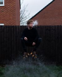 Full length of young man levitating while smoking against fence