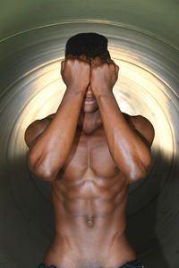 Shirtless young man with hands covering face standing against wall