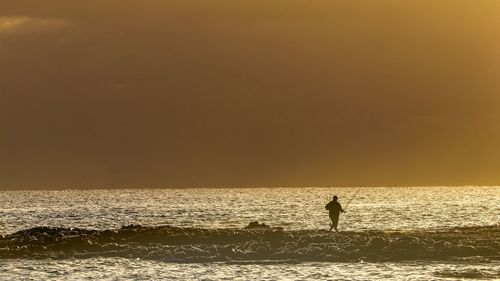 Silhouette person standing in sea against sky during sunset
