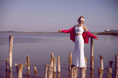 Portrait of young woman in pink and white clothes wearing sunglasses stands on stump on pink lake 