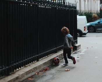 Rear view of boy playing with soccer on street by fence