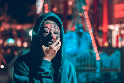 Young man wearing mask standing against illuminated lights at night
