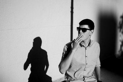 Young man wearing sunglasses smoking cigarette against wall during sunny day