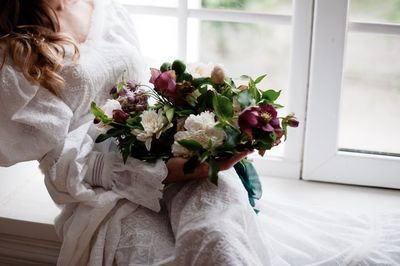 Midsection of woman holding flowers while sitting on window sill
