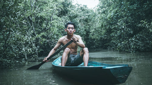 Portrait of shirtless man sitting in lake against trees